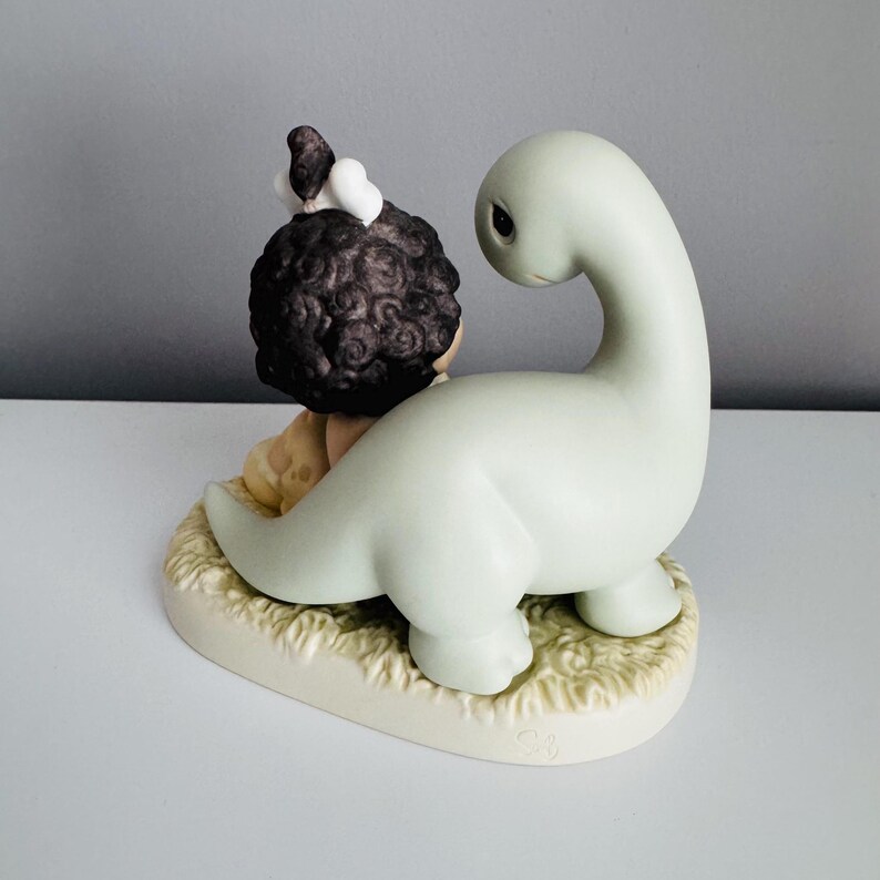 Może przedstawiać: Ceramiczna figurka przedstawiająca dziecko z ciemnymi kręconymi włosami i małym ptakiem, obok jasnozielonego dinozaura. Figurki umieszczone są na podstawie o słomianej fakturze, tworząc kapryśną scenę.