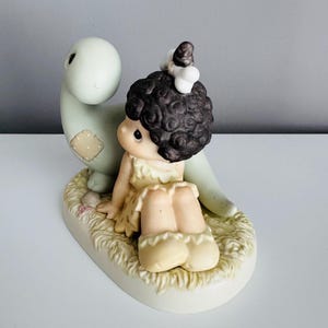 Może przedstawiać: Ceramiczna figurka przedstawiająca dziewczynkę z ciemnymi kręconymi włosami i jasnoż&oacute;łtą sukienką, siedzącą obok jasnozielonego dinozaura. Owalna podstawa ma fakturę przypominającą słomę. Figurka ma miękką, pastelową paletę kolor&oacute;w.