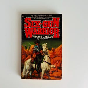 Pu&ograve; includere: Libro tascabile vintage intitolato "Sex-Gun Warrior" di Prairie Caesar. La copertina presenta un cowboy su un cavallo bianco, una donna e il titolo in lettere rosse in grassetto. Lo sfondo &egrave; una scena desertica.