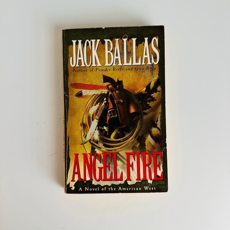 Pu&ograve; includere: Un libro tascabile intitolato "ANGEL FIRE" di Jack Ballas. La copertina presenta un tema western con una pistola, un coltello e una corda. &Egrave; visibile anche il testo "A Novel of the American West".