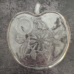 May include: A clear glass serving plate shaped like an apple with a stem at the top. The center of the plate features an embossed design of a flower with petals, leaves, and a small circular ring detail positioned above the flower.