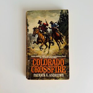 Pu&ograve; includere: Libro tascabile vintage intitolato "Colorado Crossfire" di Patrick E. Andrews. La copertina raffigura due cowboy a cavallo, con le pistole estratte, in una scena western. Il titolo del libro &egrave; in grandi lettere arancioni.
