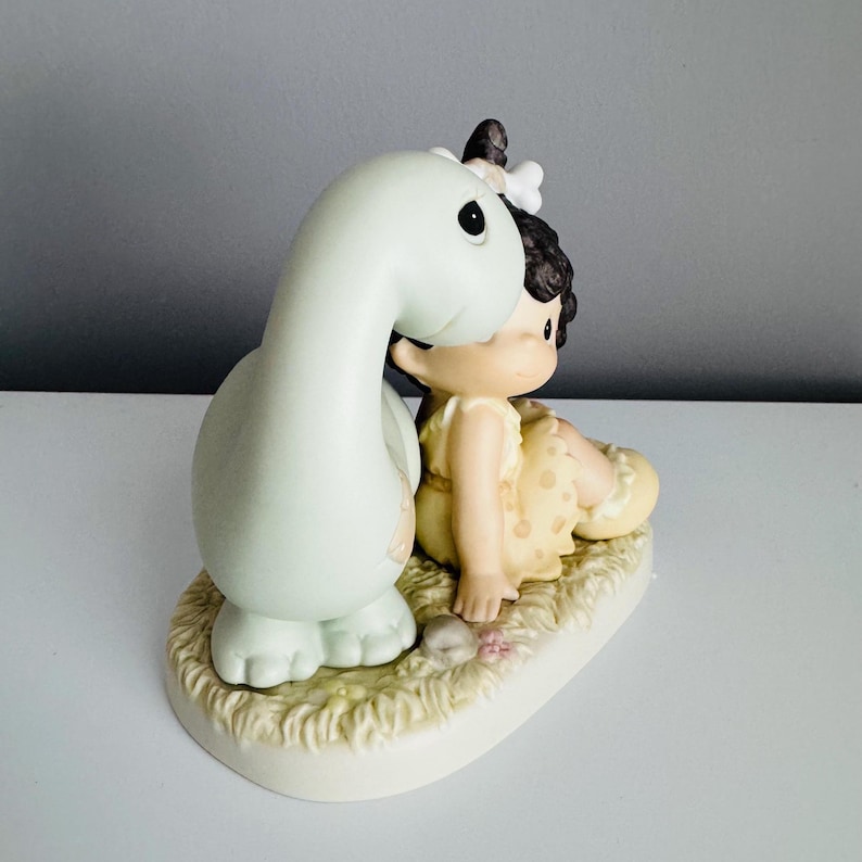Może przedstawiać: Ceramiczna figurka przedstawiająca jasnozielonego dinozaura i siedzące dziecko w ż&oacute;łtej sukience z brązowymi włosami. Figurka umieszczona jest na podstawie o trawiastym wzorze i małych kamieniach.