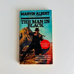 Pu&ograve; includere: Un libro tascabile vintage intitolato "THE MAN IN BLACK" di Marvin Albert. La copertina presenta un uomo in nero con una pistola, ambientato su uno sfondo desertico. Il libro &egrave; etichettato come un Gold Medal Classic Western.