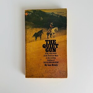 Pu&ograve; includere: Libro tascabile vintage intitolato "The Quiet Gun" di Leo Brady. La copertina presenta una scena western con un cowboy a cavallo e due cavalli. Il libro ha una copertina gialla e marrone con il testo "A big, lusty novel of the American West."