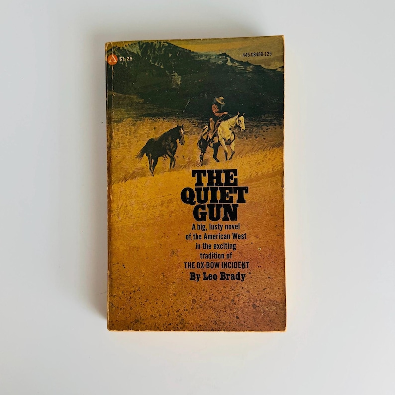 Pu&ograve; includere: Libro tascabile vintage intitolato "The Quiet Gun" di Leo Brady. La copertina presenta una scena western con un cowboy a cavallo e due cavalli. Il libro ha una copertina gialla e marrone con il testo "A big, lusty novel of the American West."