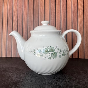May include: A white ceramic teapot with a rounded body and a subtle spiral texture. The teapot features a green ivy leaf pattern around the center, a matching lid with a small knob handle, a curved spout, and a sturdy handle on the right side.