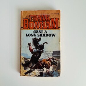 Pu&ograve; includere: Libro tascabile vintage intitolato "Cast a Long Shadow" di Frank Bonham. La copertina presenta un cowboy su un cavallo che si impenna, con un secondo cowboy a terra. Il libro ha uno schema di colori arancione e marrone.