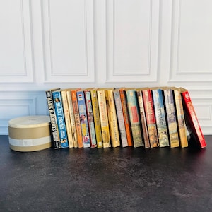 May include: A collection of vintage books with colorful spines, arranged on a dark surface. Titles include "Mark K. Roberts", "Silent Drums", and "Springfield 45/70". A round, light-colored box sits to the left of the books.