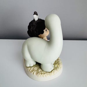 Może przedstawiać: Ceramiczna figurka dziecka obejmującego jasnozielonego dinozaura. Dziecko ma ciemne kręcone włosy i ma na sobie jasny str&oacute;j. Mały ptaszek siedzi na głowie dziecka. Podstawa jest w kolorze złamanej bieli o fakturze trawy.