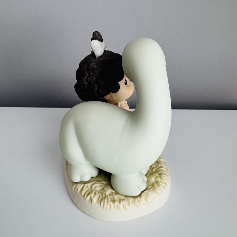 Może przedstawiać: Ceramiczna figurka dziecka obejmującego jasnozielonego dinozaura. Dziecko ma ciemne kręcone włosy i ma na sobie jasny str&oacute;j. Mały ptaszek siedzi na głowie dziecka. Podstawa jest w kolorze złamanej bieli o fakturze trawy.