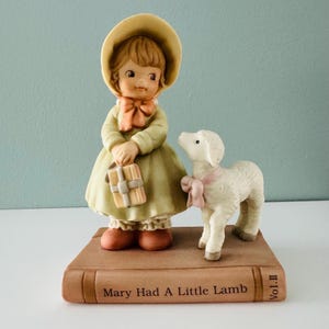 May include: A ceramic figurine of a girl in a yellow hat and dress, holding a gift, with a lamb wearing a pink bow. The figurine is on a book-shaped base that reads "Mary Had A Little Lamb".
