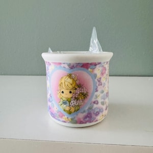 May include: A ceramic mug with a floral pattern in shades of pink, purple, and yellow. The mug features a heart-shaped design with a cartoon illustration of a girl holding flowers and a book. A plastic wrapper is inside the mug.