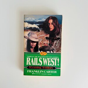 Pu&ograve; includere: Un libro tascabile intitolato "RAILS WEST! Wyoming Territory" di Franklin Carter. La copertina presenta illustrazioni di persone, un treno e un bisonte sullo sfondo di montagne. Il libro ha un bordo verde e bianco.