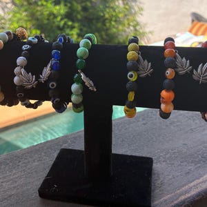 May include: A display of beaded bracelets in various colors, including green, blue, orange, and red. Each bracelet features a silver leaf charm and a mix of stone and glass beads. Some bracelets include evil eye beads.