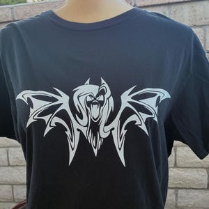 May include: Black t-shirt featuring a white graphic of a stylized bat with open wings and a snarling face. The design is centered on the front of the shirt. The shirt has short sleeves and a crew neck.