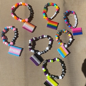 May include: A collection of eight colorful beaded bracelets with charms. Each bracelet features a different pride flag and the words "TRANS", "ACE", "NON-BINARY", "LESBIAN", "BISEXUAL", "PAN", and "TRANS".