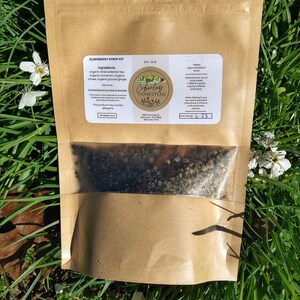 May include: A brown paper bag with a clear window showing a mixture of dried herbs and spices. The bag is labeled "Elderberry Syrup Kit" and includes the ingredients: organic dried elderberries, organic cinnamon, organic cloves, organic ground ginger, and honey. The bag also includes instructions for making the syrup and a note about potential allergens.