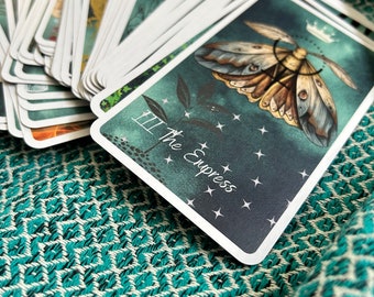 May include: A deck of tarot cards with a teal and white patterned background. The top card features a moth illustration with the words "III The Empress" and star accents. The cards are fanned out, revealing other illustrated cards.