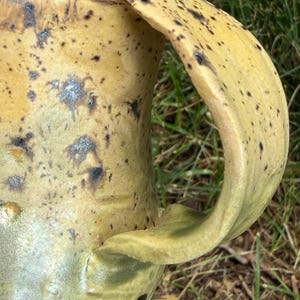 May include: A ceramic mug featuring a yellow glaze with dark brown speckles and subtle green undertones. The mug has a wide, curved handle attached to the side, with a smooth, uneven surface texture and a visible rim at the top of the vessel.