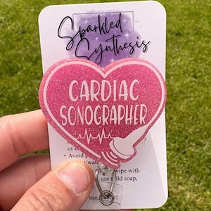 Peut inclure: Un badge rose pailleté en forme de cœur avec les mots "CARDIAC SONOGRAPHER" en blanc. Le badge présente également une image d'échographie médicale et le texte "Sparkled Synthesis". Le badge est fixé à un enrouleur rétractable.