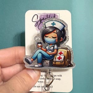 以下が含まれることがあります： マスクをした看護師の漫画のキャラクターがコーヒーカップを持ち、赤い十字の付いたバックパックの隣に座っているバッジリール。 バッジリールはキラキラした縁取りと透明な格納式コードが付いています。
