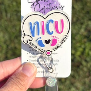 Może przedstawiać: Uchwyt na identyfikator z sercowatym wzorem z literami "NICU" w kolorze niebieskim i różowym, ze stetoskopem, odciskami stóp i tekstem "Where Little Things Matter". Uchwyt na identyfikator jest przymocowany do przezroczystego plastikowego uchwytu.