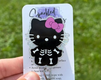 Pode incluir: Um porta-crachá em forma de gato preto com um laço rosa e uma estampa branca de caixa torácica esquelética. O personagem tem bigodes e olhos brancos sobre um fundo brilhante em um cartão branco com o texto Sparkled no topo.