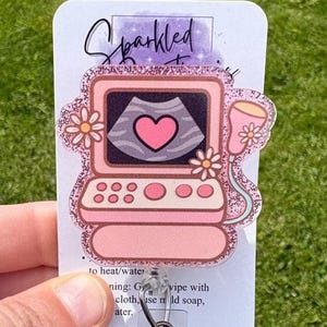 May include: A pink and glittery badge reel featuring an ultrasound machine design with a heart-shaped image on the screen. The badge reel has a clear plastic clip and a retractable cord. The word "Sparkled" is written on the top of the badge reel.