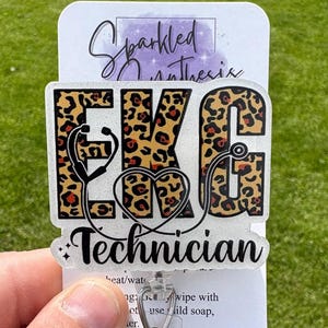 May include: A badge reel with the words "EKG Technician" in black lettering. The letters "EKG" are in a leopard print pattern. A stethoscope graphic is included. The badge reel is attached to a clear plastic holder with a glittery, round embellishment.