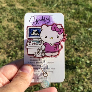 May include: A sparkly pink Hello Kitty badge reel with a pink bow and a purple shirt. The reel features a small ultrasound image and the text "Sparkled". The back of the reel has instructions for cleaning and care.