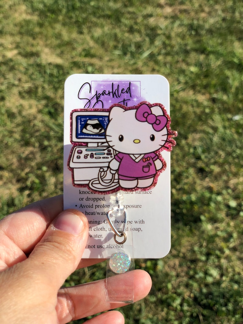 May include: A sparkly pink Hello Kitty badge reel with a pink bow and a purple shirt. The reel features a small ultrasound image and the text "Sparkled". The back of the reel has instructions for cleaning and care.
