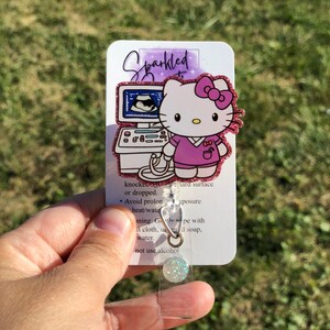 May include: A pink glitter Hello Kitty badge holder with a pink and white ultrasound image. The badge holder has a clear plastic cover and a silver metal clip. The text on the badge holder reads "Sparkled" and "knocked or dropped. Avoid prolonged heat/water exposure. Gently wipe with a soft cloth, using mild soap and water. Do not use alcohol."