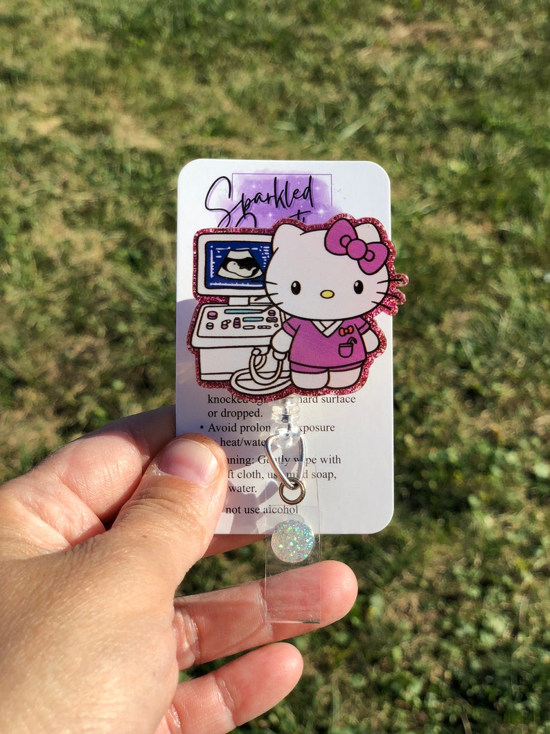 May include: A pink glitter Hello Kitty badge holder with a pink and white ultrasound image. The badge holder has a clear plastic cover and a silver metal clip. The text on the badge holder reads "Sparkled" and "knocked or dropped. Avoid prolonged heat/water exposure. Gently wipe with a soft cloth, using mild soap and water. Do not use alcohol."