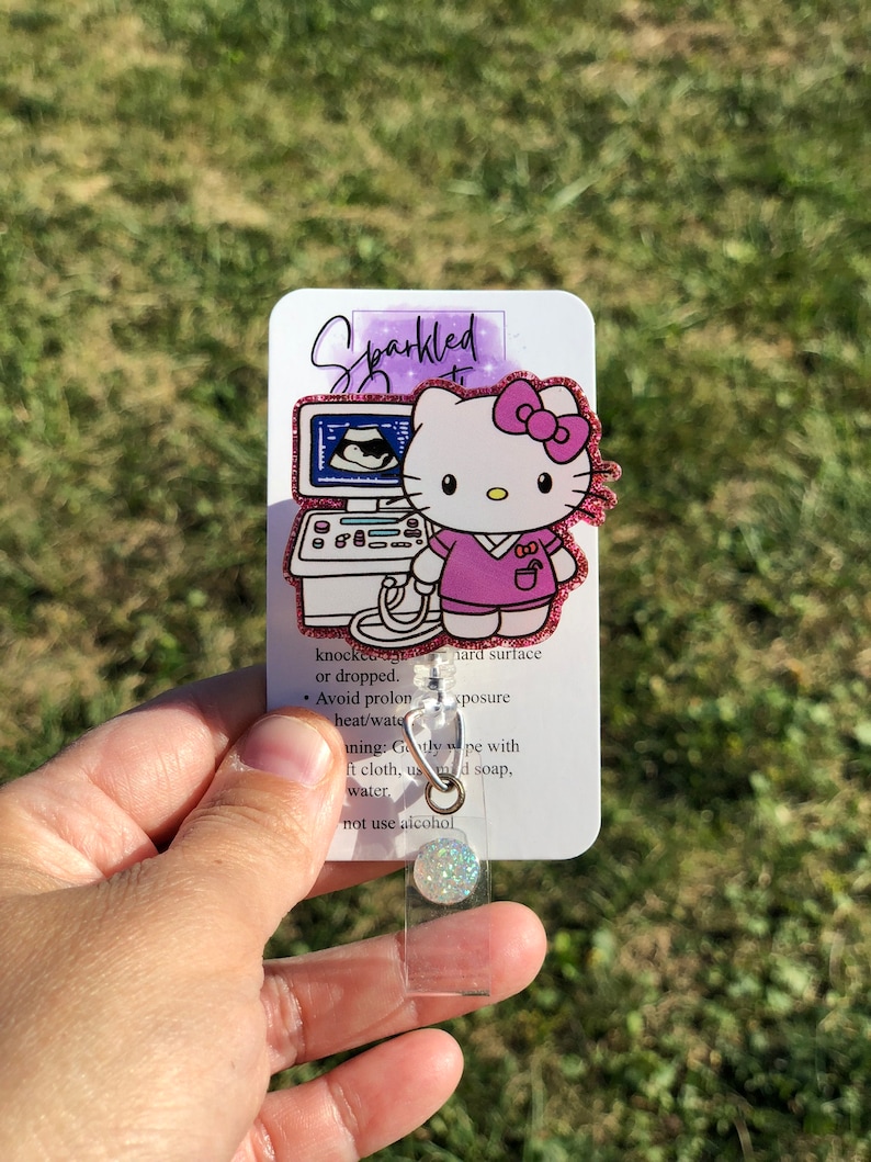 May include: Pink glitter Hello Kitty ultrasound technician retractable badge reel.  The reel features Hello Kitty in a pink scrub top, in front of an ultrasound machine. The card attached includes care instructions: Avoid prolonged heat/water exposure; Gently wipe with a soft cloth, mild soap, and water; Do not use alcohol.  The text 'Sparkled' is visible on the card.