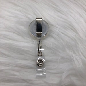 May include: A clear plastic retractable badge holder with a silver clip and a silver swivel hook. The holder has a clear plastic strap with a silver circle on it.