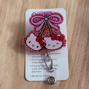May include: Pink Hello Kitty charm with a pink bow and cherries. The charm is attached to a clear plastic retractable badge holder with a silver metal clip.