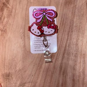 May include: Pink glitter Hello Kitty charm with a pink bow and a cherry design. The charm is attached to a clear plastic retractable badge holder.  The back of the charm has instructions for cleaning and care.  "Knocked or dropped. Avoid prolonged exposure to heat/water. Cleaning: Gently wipe with a soft cloth, use mild soap and water. Do not use alcohol."