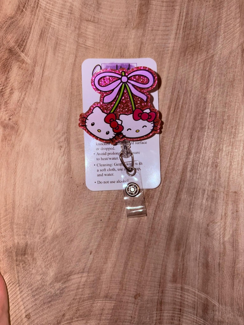 May include: Pink glitter Hello Kitty charm with a pink bow and a cherry design. The charm is attached to a clear plastic retractable badge holder.  The back of the charm has instructions for cleaning and care.  "Knocked or dropped. Avoid prolonged exposure to heat/water. Cleaning: Gently wipe with a soft cloth, use mild soap and water. Do not use alcohol."