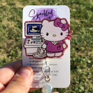 May include: A white and purple Hello Kitty badge reel with a pink bow and a purple scrub top. The badge reel features a cartoon image of Hello Kitty holding an ultrasound machine. The text "Sparkled" is written on the card. The card also includes instructions for cleaning and care.