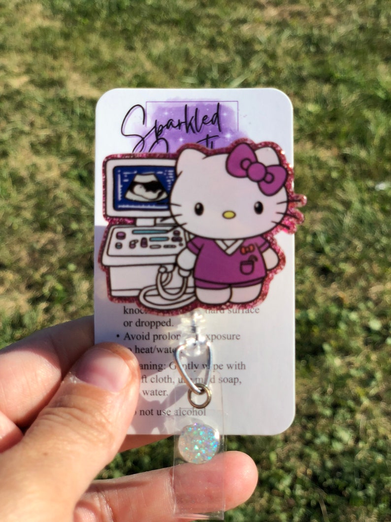 May include: A white and purple Hello Kitty badge reel with a pink bow and a purple scrub top. The badge reel features a cartoon image of Hello Kitty holding an ultrasound machine. The text "Sparkled" is written on the card. The card also includes instructions for cleaning and care.