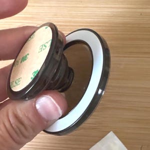 Puede incluir: Un soporte circular negro para teléfono con base extensible tipo acordeón y un adhesivo trasero circular con texto 3M en verde. El dispositivo incluye un componente de anillo negro secundario con un borde interior blanco para soporte.