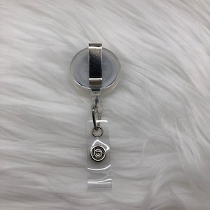 May include: A clear retractable badge holder with a silver clip and a silver circle with a design in the center.