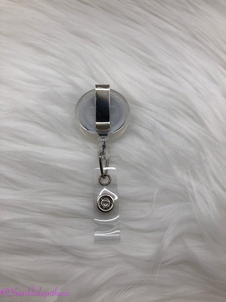 May include: A clear retractable badge holder with a silver clip and a silver circle with a design in the center.