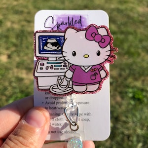 May include: A pink and white Hello Kitty character with a purple shirt and a stethoscope. The character is standing in front of an ultrasound machine. The image is on a white card with the text "Sparkled" and instructions for care. The card is attached to a clear retractable badge holder with a sparkly blue and white button.