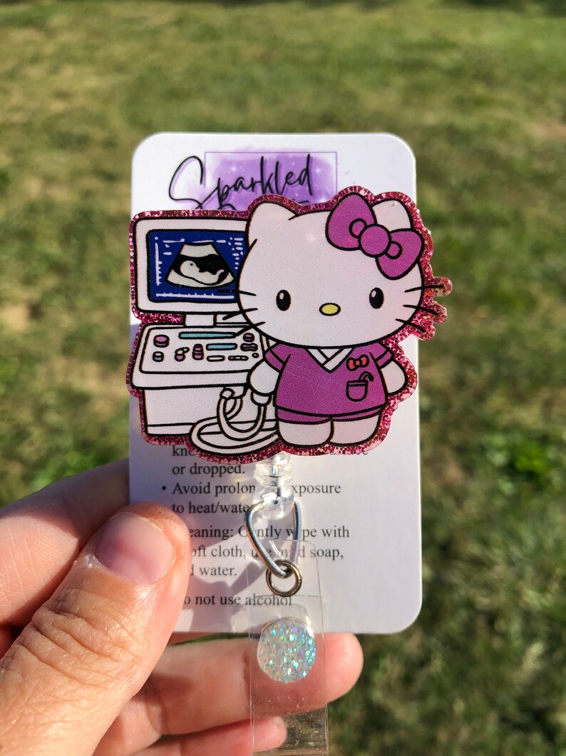 May include: A pink and white Hello Kitty character with a purple shirt and a stethoscope. The character is standing in front of an ultrasound machine. The image is on a white card with the text "Sparkled" and instructions for care. The card is attached to a clear retractable badge holder with a sparkly blue and white button.