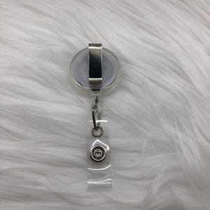 May include: A clear retractable badge holder with a silver clip and a silver circle with a design in the center.