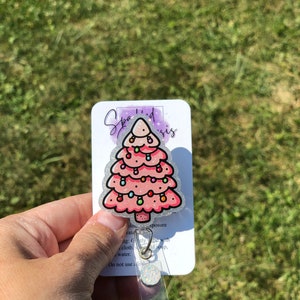 May include: A pink glitter Christmas tree with white lights and ornaments. The tree is on a white card with the text "Sparkled Sis" and instructions for care. The card is attached to a clear plastic badge holder.