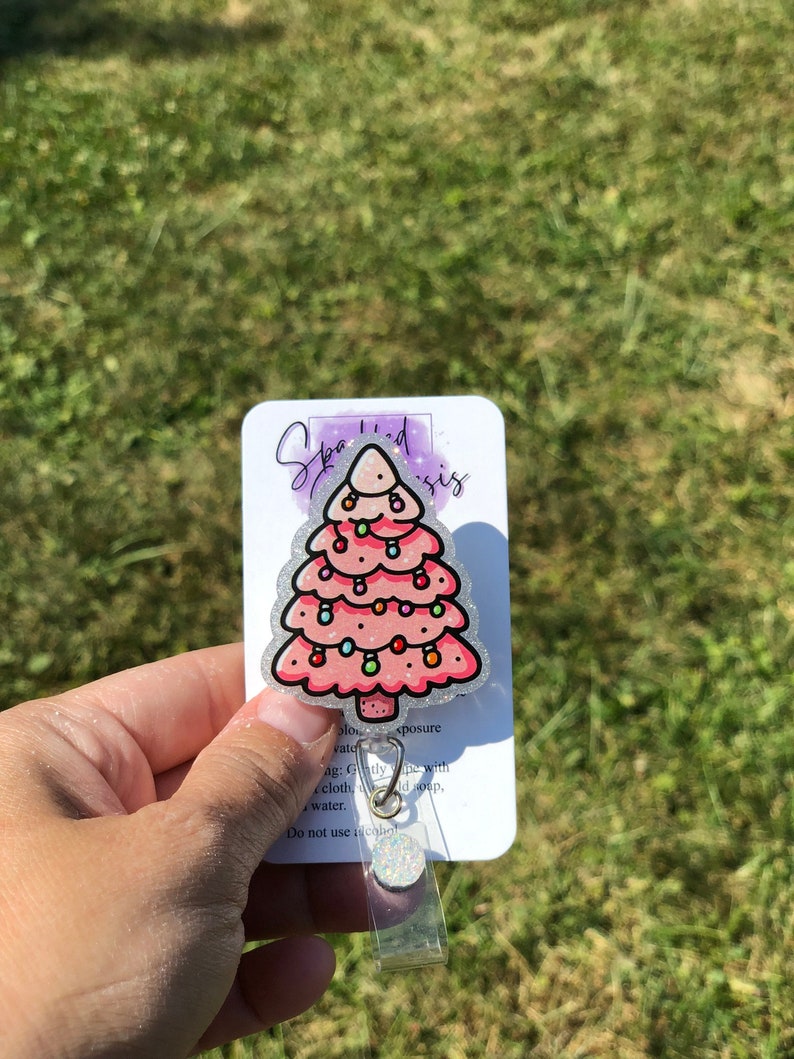 May include: A pink glitter Christmas tree with white lights and ornaments. The tree is on a white card with the text "Sparkled Sis" and instructions for care. The card is attached to a clear plastic badge holder.