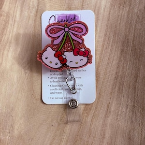 May include: Pink glitter Hello Kitty charm with a pink bow and cherries. The charm is attached to a clear plastic badge holder with a metal clip. The card behind the charm has instructions for care: "knocked against a hard surface or dropped. Avoid prolonged exposure to heat/water. Cleaning: Gently wipe with a soft cloth, use soap, and water. Do not use alcohol."